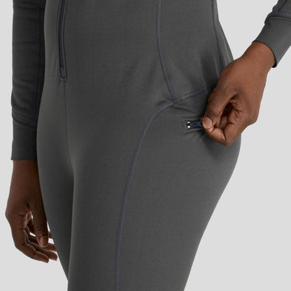 A woman wearing the NRS Expedition Weight Union Suit, featuring a gray fleece design with a zipper pocket, is shown. Her hand grips the zipper, highlighting its sustainable construction from recycled materials.