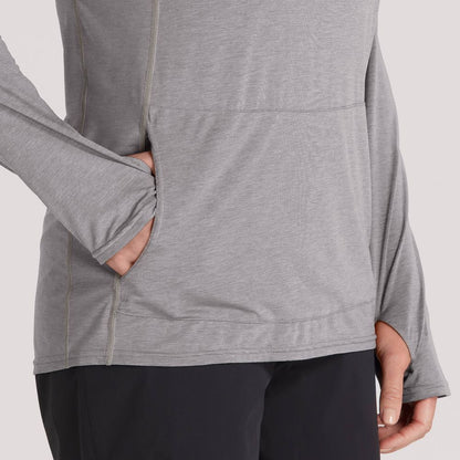 A person in an NRS Silkweight Vesi Hoodie - Women's, gray long-sleeve made from recycled material, black pants, and one hand in the hoodie pocket stands against a plain background.