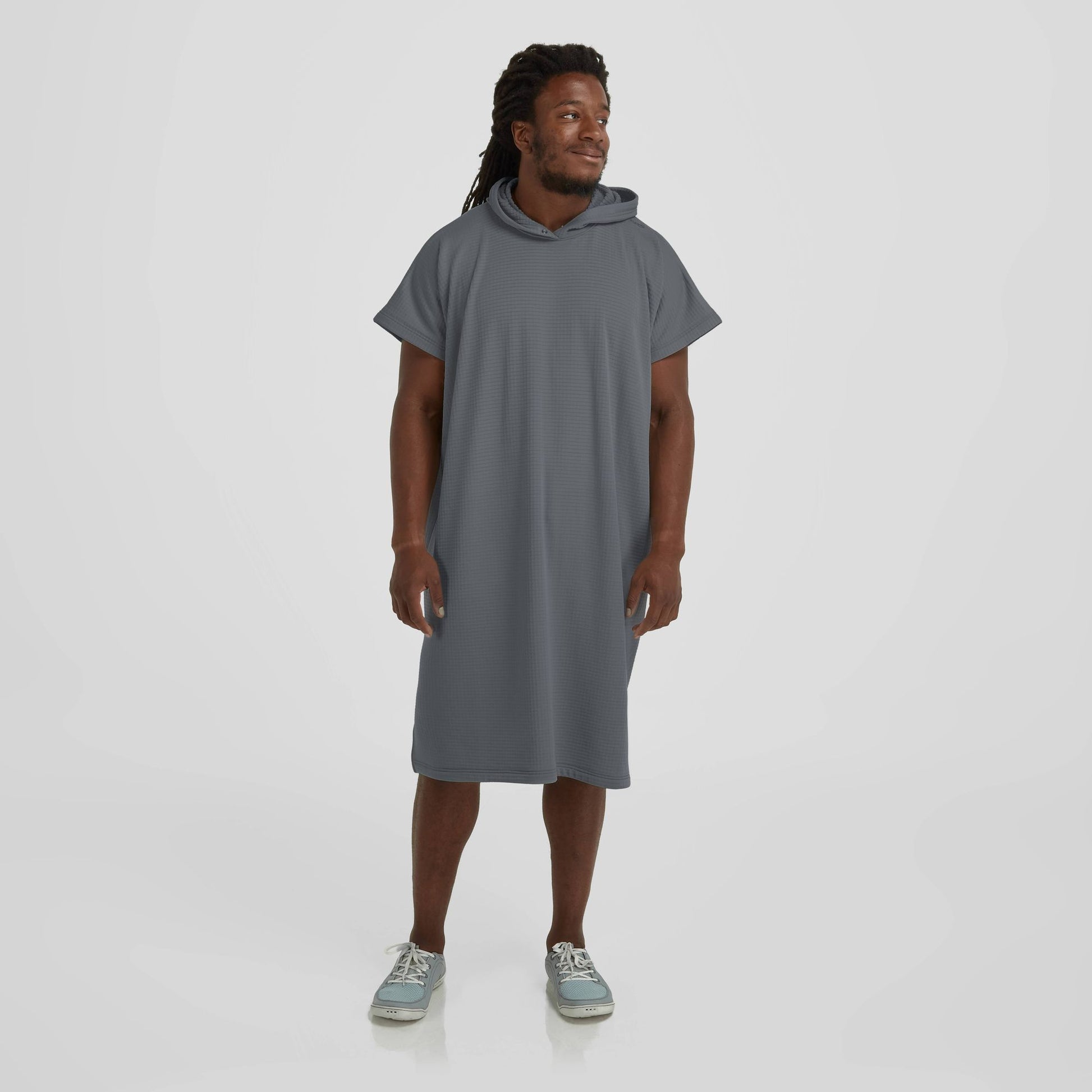 A man with long hair stands against a plain background, wearing a short-sleeve gray NRS Covert Changing Poncho made of lightweight fabric and casual shoes. He is looking off to the side and smiling slightly.