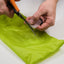 Someone uses orange-handled scissors to cut green tissue paper on a white surface, like how Gear Aid’s Tenacious Tape Silnylon Repair Tape is trimmed to patch ripstop nylon.