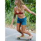 Amidst lush greenery, a person with long hair skateboards barefoot on a path, wearing a red bikini top and Carve's Oahu 4" Short in blue stretch cotton corduroy. It's the perfect addition to any vacation wardrobe.