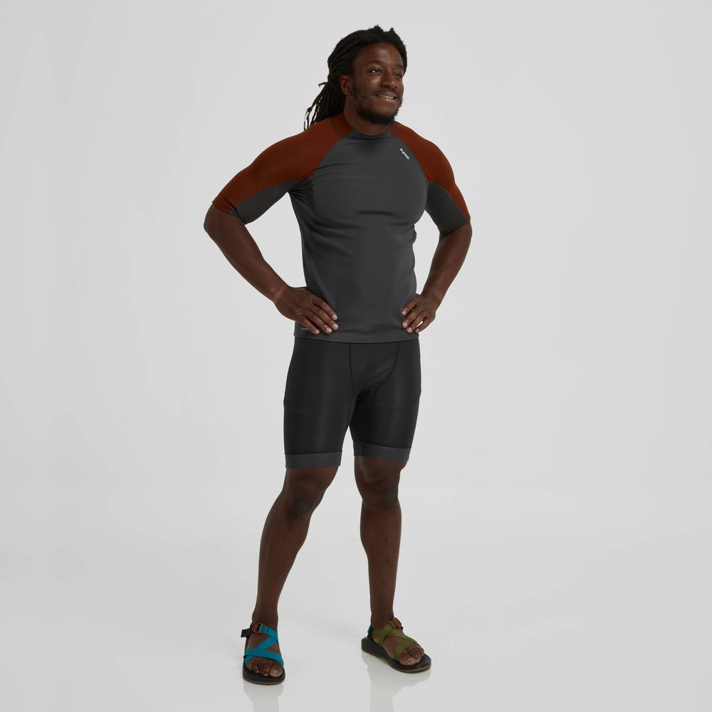 A man confidently stands against a plain background wearing an NRS Hydroskin 0.5mm Short Sleeve Shirt in dark gray and red, ideal for paddling performance, paired with black shorts and sandals.