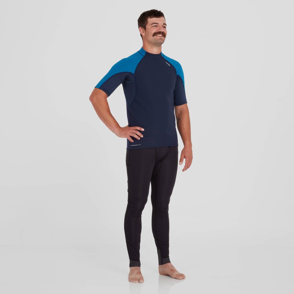 A man stands barefoot in a navy NRS Hydroskin 0.5mm Short Sleeve Shirt with blue shoulders and black leggings, boosting his paddling performance against a plain white backdrop.
