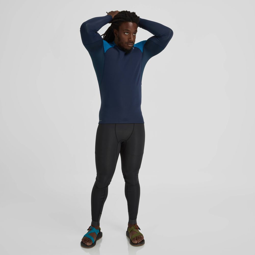 Person wearing a NRS Hydroskin 0.5mm Long Sleeve Shirt in navy, black athletic leggings, and teal and brown sandals, standing against a plain white background.