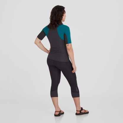 A woman wearing an NRS HydroSkin 0.5mm Short Sleeve Shirt in dark and teal, paired with sandals, stands with her back to the camera against a plain background.