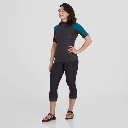 A person stands against a plain background wearing the NRS Hydroskin 0.5mm Short Sleeve Shirt and capri pants with sandals, an outfit perfect for minimizing water retention.
