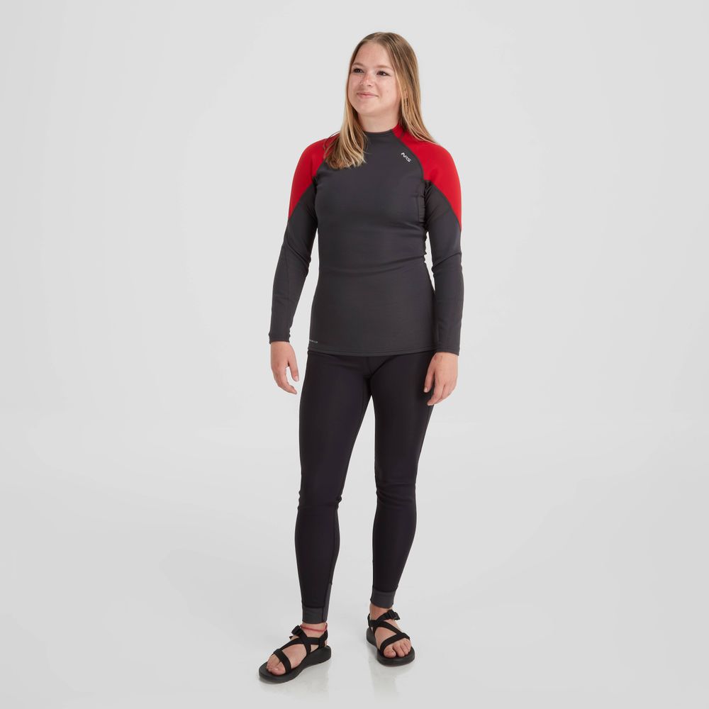 A person wearing a black and red NRS Women's Hydroskin 0.5mm Long Sleeve Shirt with black leggings and sandals stands against a plain white background, ready to enhance paddling performance.