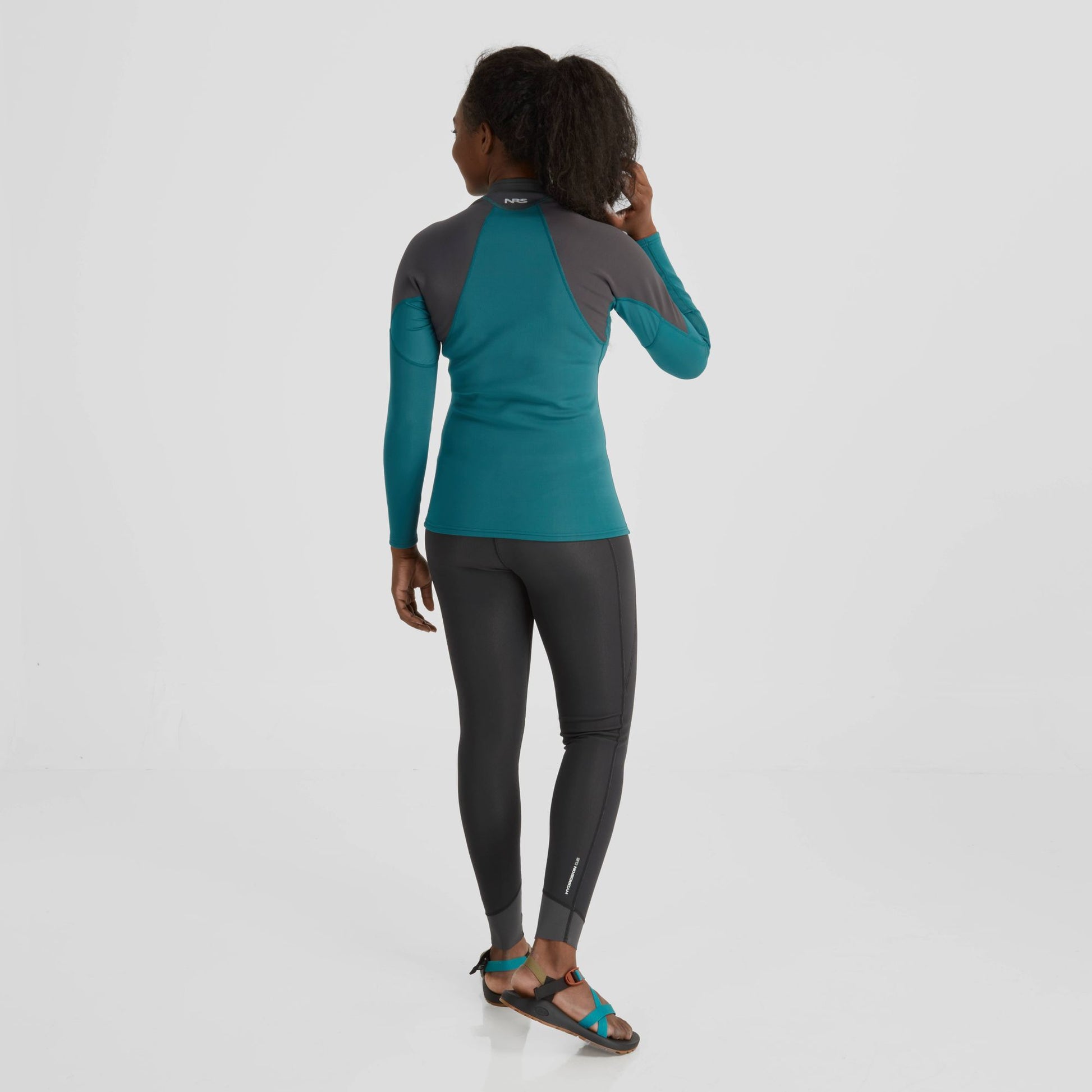 The back view of a woman wearing the NRS Hydroskin 0.5 Long Sleeve Shirt - Women's, an insulating top, and leggings.