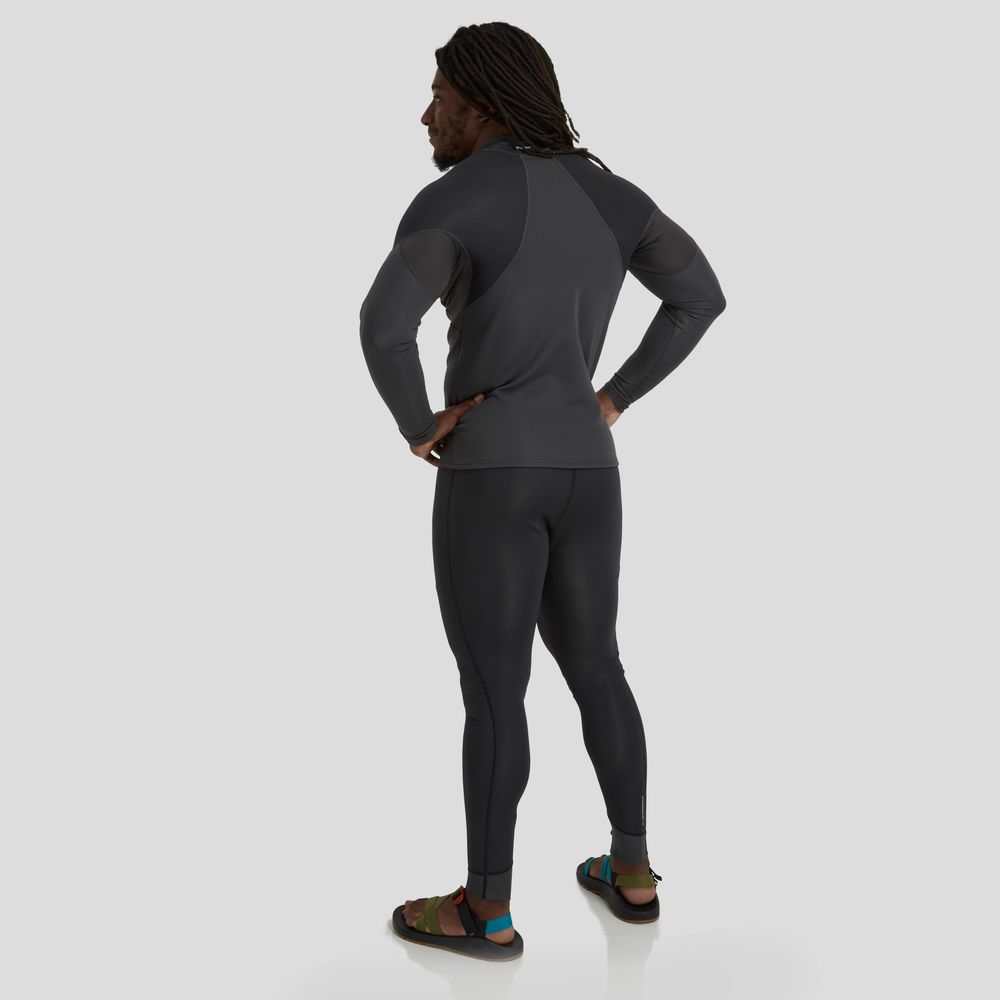 A person stands with hands on hips, sporting long braided hair and sandals while facing away. They're dressed in a dark athletic outfit, complemented by the NRS Hydroskin 0.5mm Jacket - Men's against a gray background.