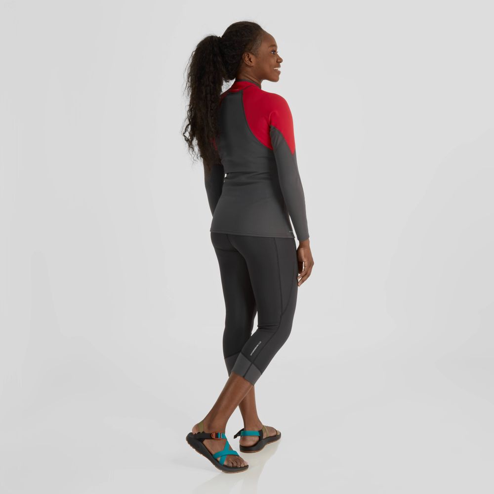 A woman in a red and gray wetsuit with black sandals is shown wearing an NRS Hydroskin 0.5mm Jacket, standing facing away while looking to her right.