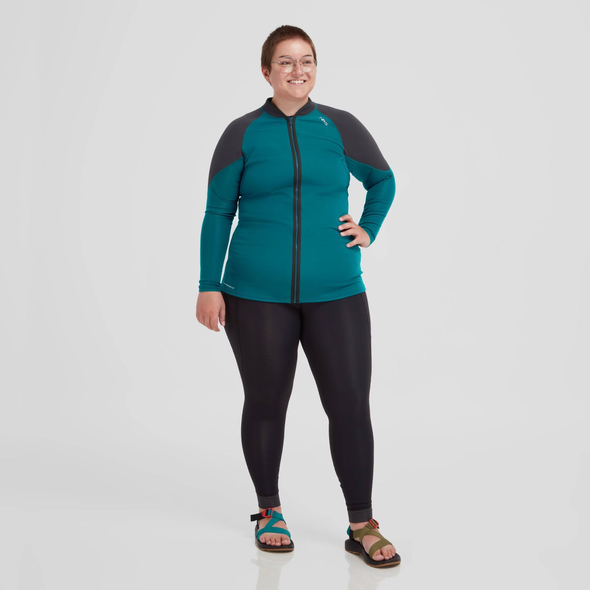A woman donning a teal and black Hydroskin 0.5 Jacket - Women's from NRS, and leggings, part of her layering arsenal for outdoor activities.