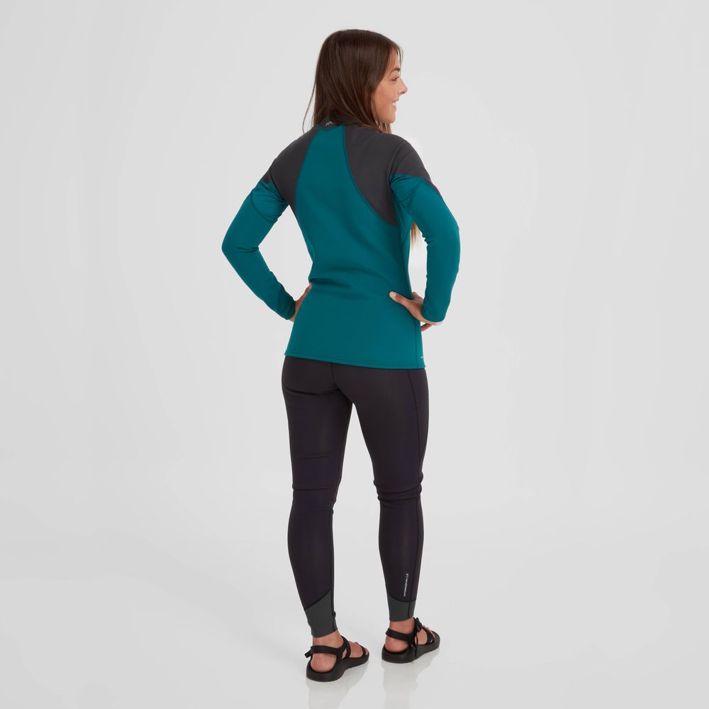 A person stands with hands on hips, wearing a teal and black long-sleeve athletic outfit similar to the NRS Hydroskin 0.5mm Jacket for women, paired with sandals against a plain white background.