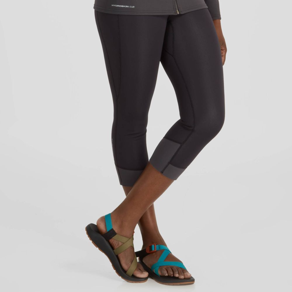 A person wearing black NRS Hydroskin 0.5mm Capri leggings and colorful sandals stands with one foot crossed over the other, showcasing paddling performance flair.