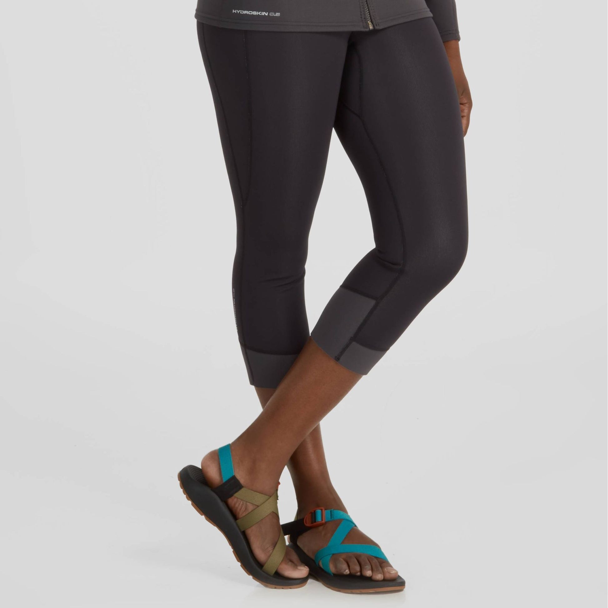 Lower body of a person wearing black NRS Hydroskin 0.5 Capri leggings and multicolored strappy sandals, standing against a plain background.