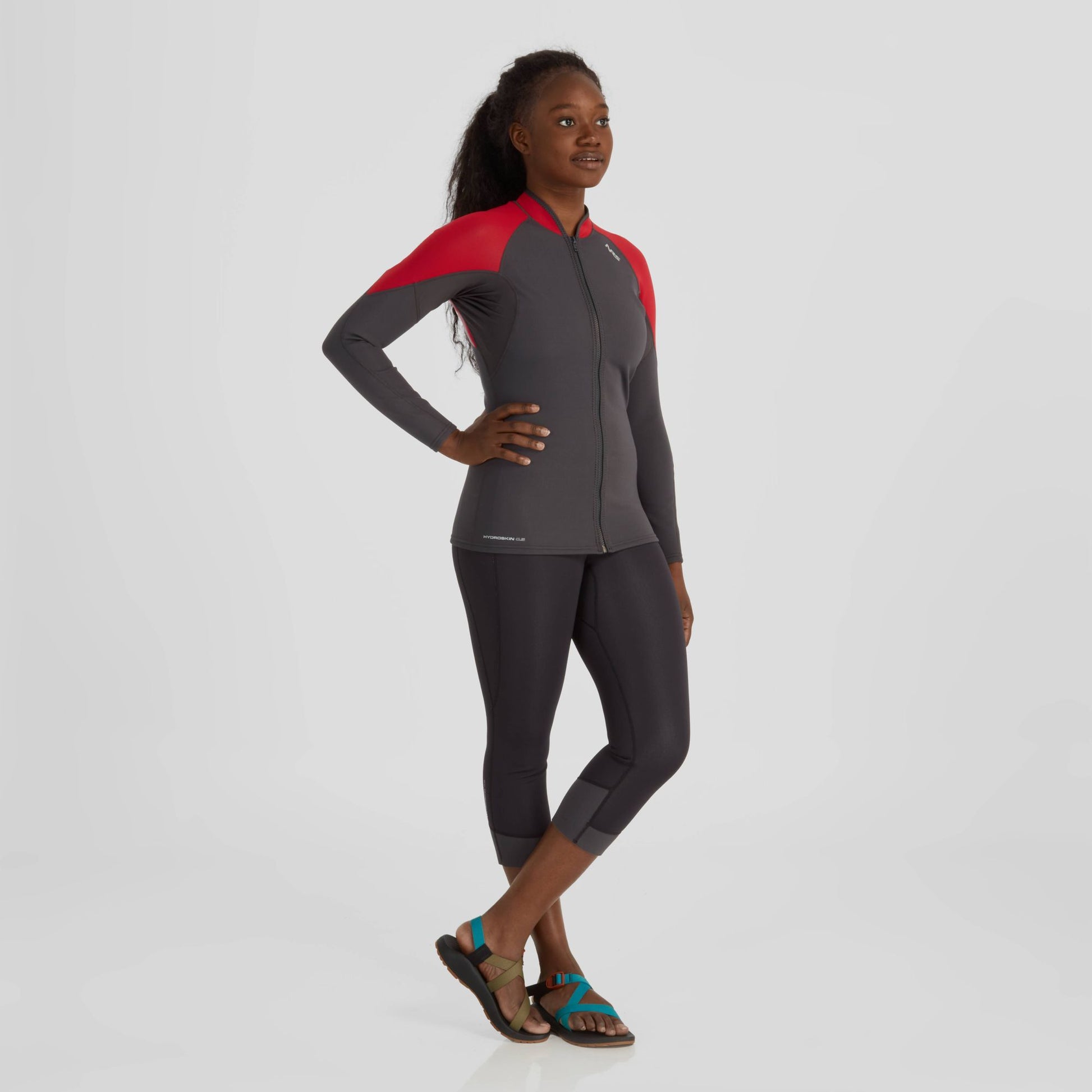 A young woman in a red and gray NRS Hydroskin 0.5 Capri wetsuit standing confidently against a white background.