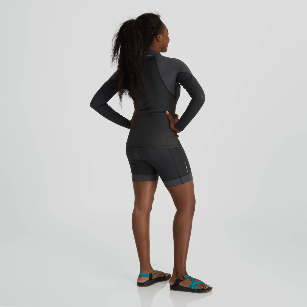 A person wearing the NRS Hydroskin 1.0mm Long Sleeve Shirt stands on a reflective surface with arms on hips, facing away. Their sleek ensemble, highlighted by neoprene protection, is complemented by sandals.