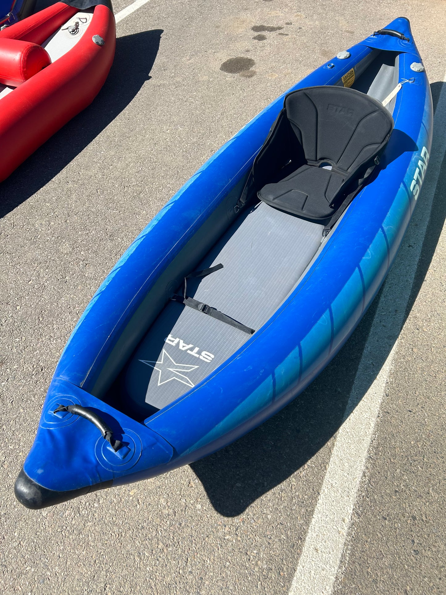 A blue Demo STAR Raven Solo inflatable kayak from NRS, featuring a black seat and gray floor pad, is parked on an asphalt surface next to a red kayak.