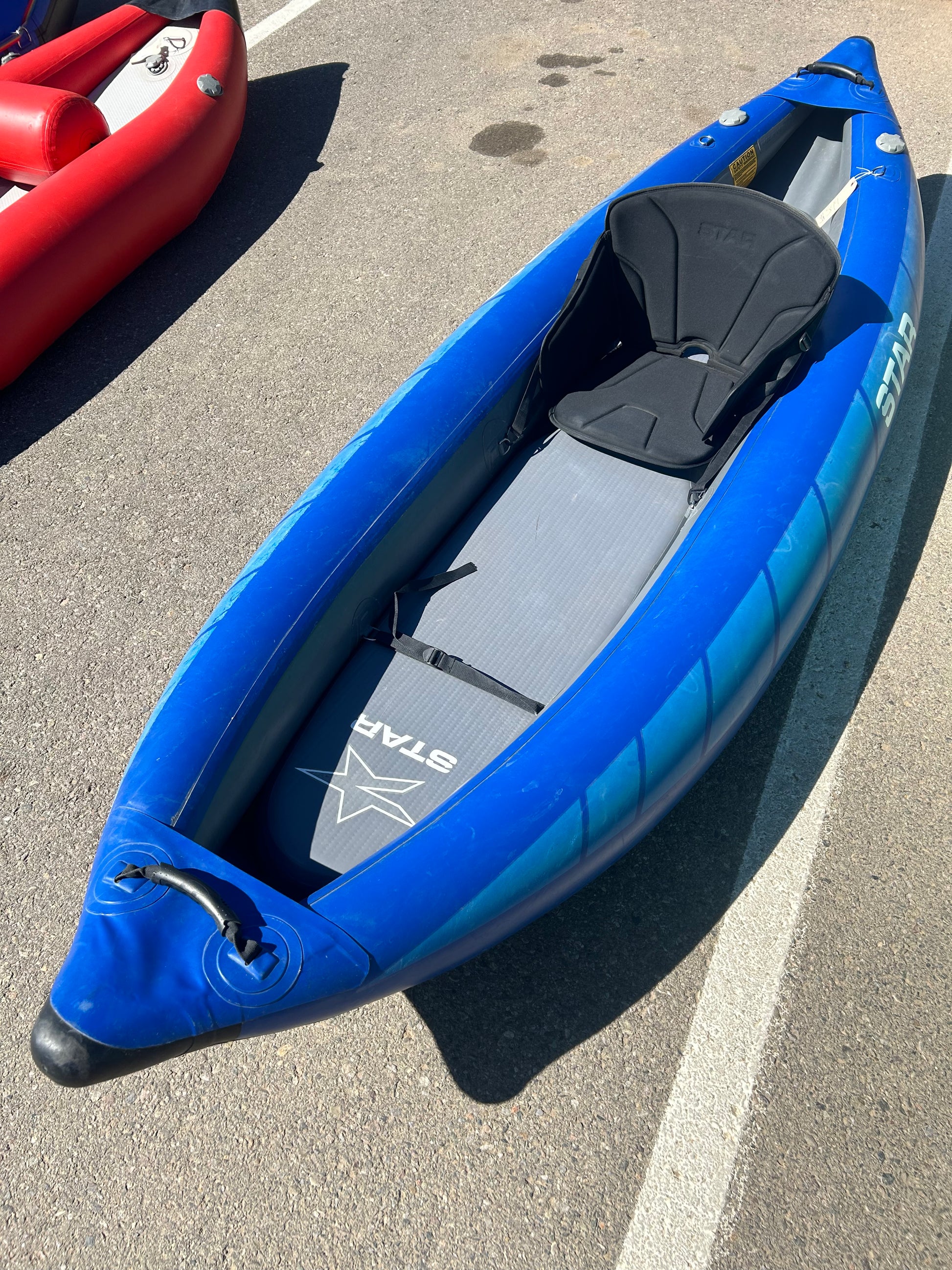 A blue Demo STAR Raven Solo inflatable kayak from NRS, featuring a black seat and gray floor pad, is parked on an asphalt surface next to a red kayak.