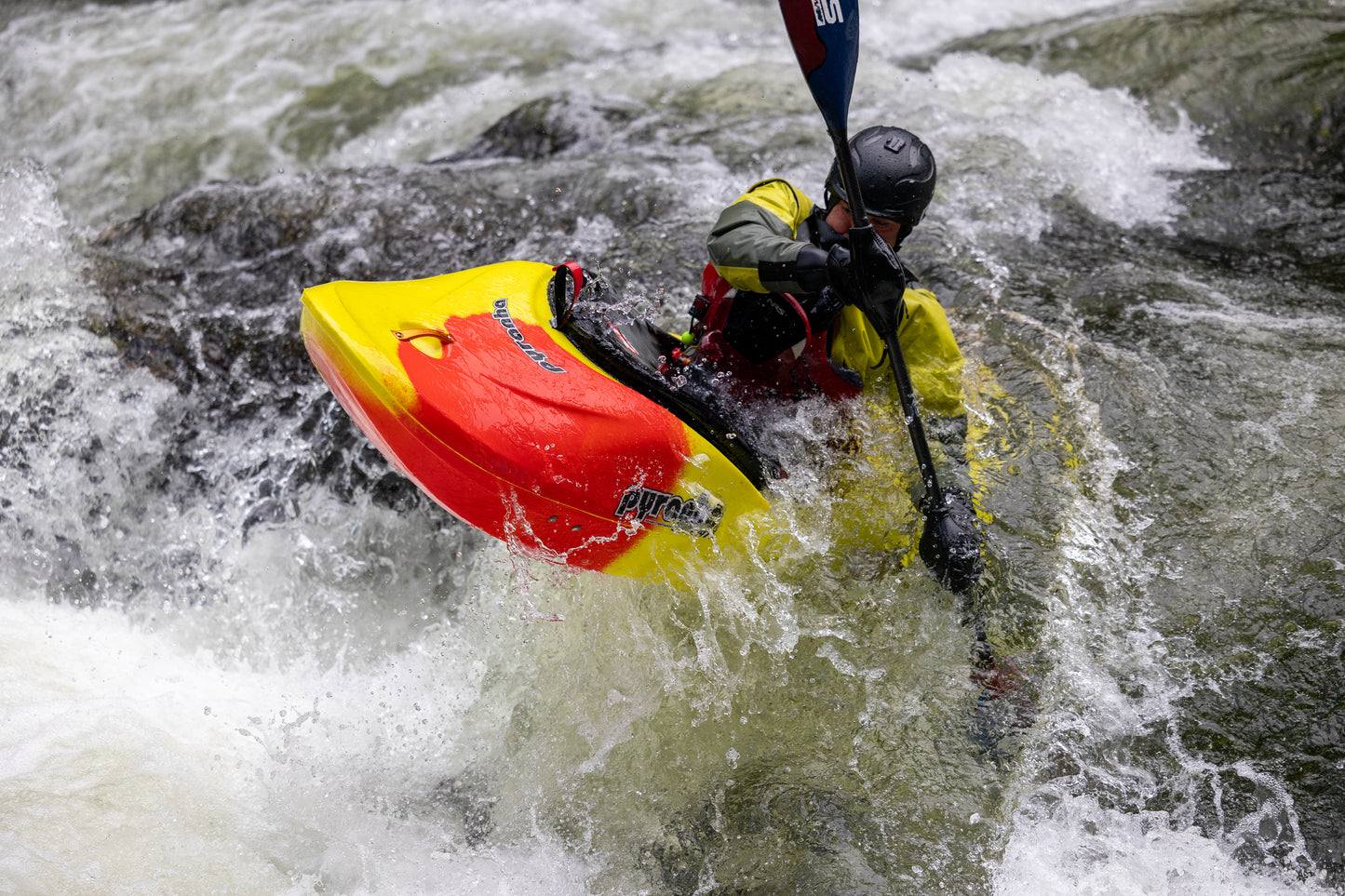 Wearing a helmet and yellow jacket, a paddler navigates rough whitewater in the Pyranha Storm kayak, demonstrating impressive downriver playboating skills.