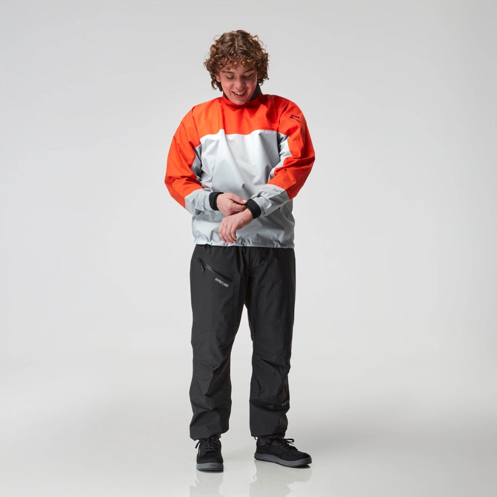 A person adjusts the sleeve of an NRS Endurance Splash Jacket for men, featuring a red and gray waterproof design, paired with black pants on a white background.