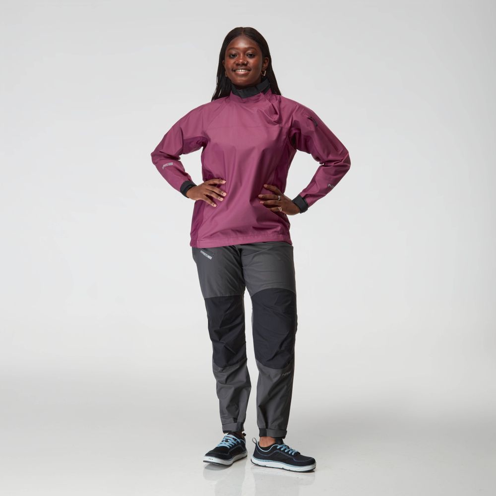 A person stands facing forward, wearing a maroon NRS Endurance Splash Jacket - Women's, gray and black pants, and black athletic shoes against a plain light background.