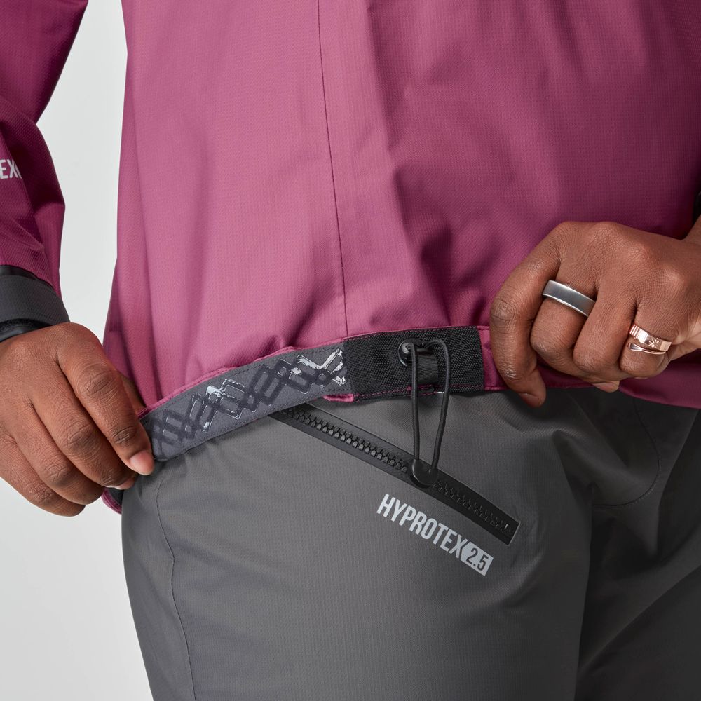 A person, torso and hand visible, pulls on the waistband of gray pants labeled "HYPROTEX 2.5" while wearing a maroon NRS Endurance Splash Jacket - Women's.