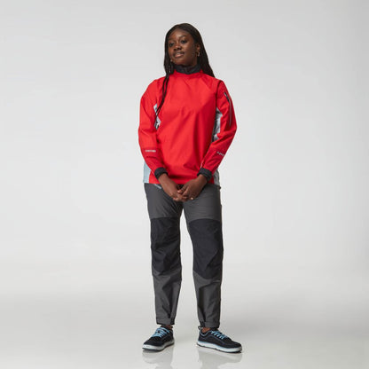 A woman stands facing the camera in an NRS Endurance Splash Jacket - Women's (red), dark gray pants, and black sneakers, set against a plain light background.