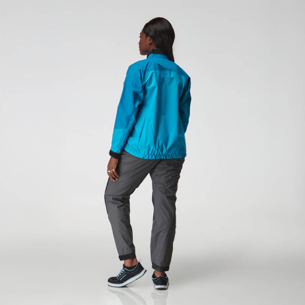 A woman faces away from the camera in a blue NRS Endurance Splash Jacket - Women's, gray pants, and blue sneakers, standing against a plain light background.