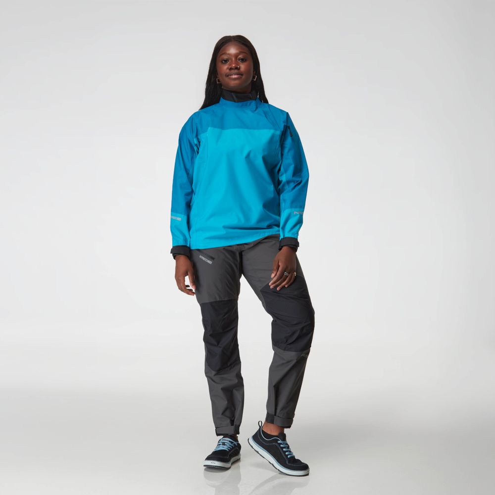 A person stands facing forward wearing the NRS Endurance Splash Jacket - Women's in bright blue, gray hiking pants, and black sneakers against a plain white background.