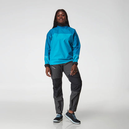 A person stands facing forward wearing the NRS Endurance Splash Jacket - Women's in bright blue, gray hiking pants, and black sneakers against a plain white background.