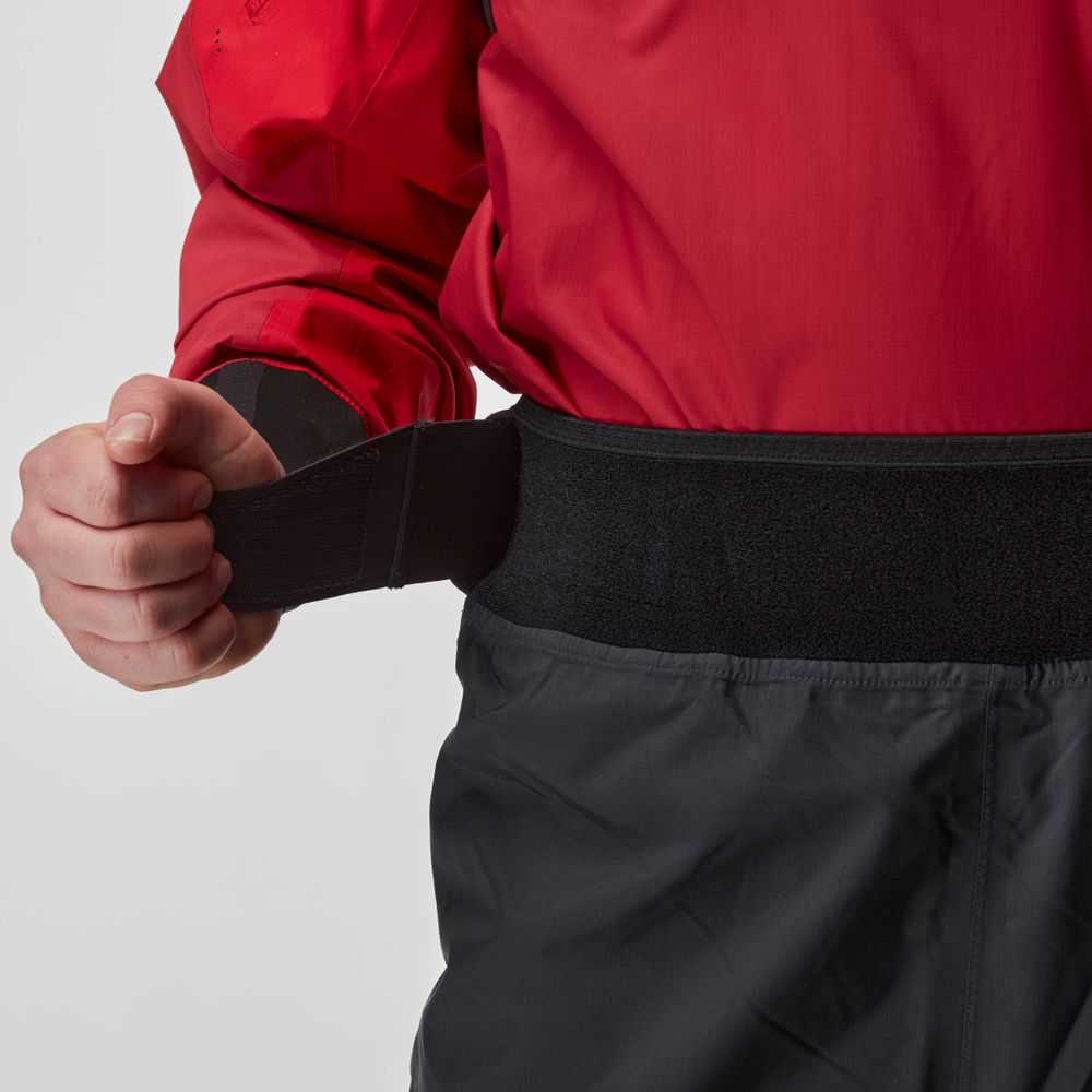 A person in a red jacket adjusts the black hook-and-loop waist strap on the NRS Endurance Splash Pant - Men's, highlighting these waterproof, breathable pants.