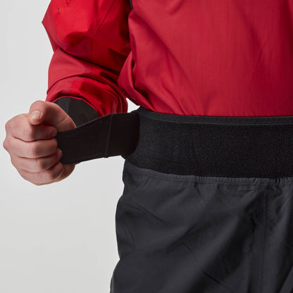 A person in a red jacket adjusts the black hook-and-loop waist strap on the NRS Endurance Splash Pant - Men's, highlighting these waterproof, breathable pants.