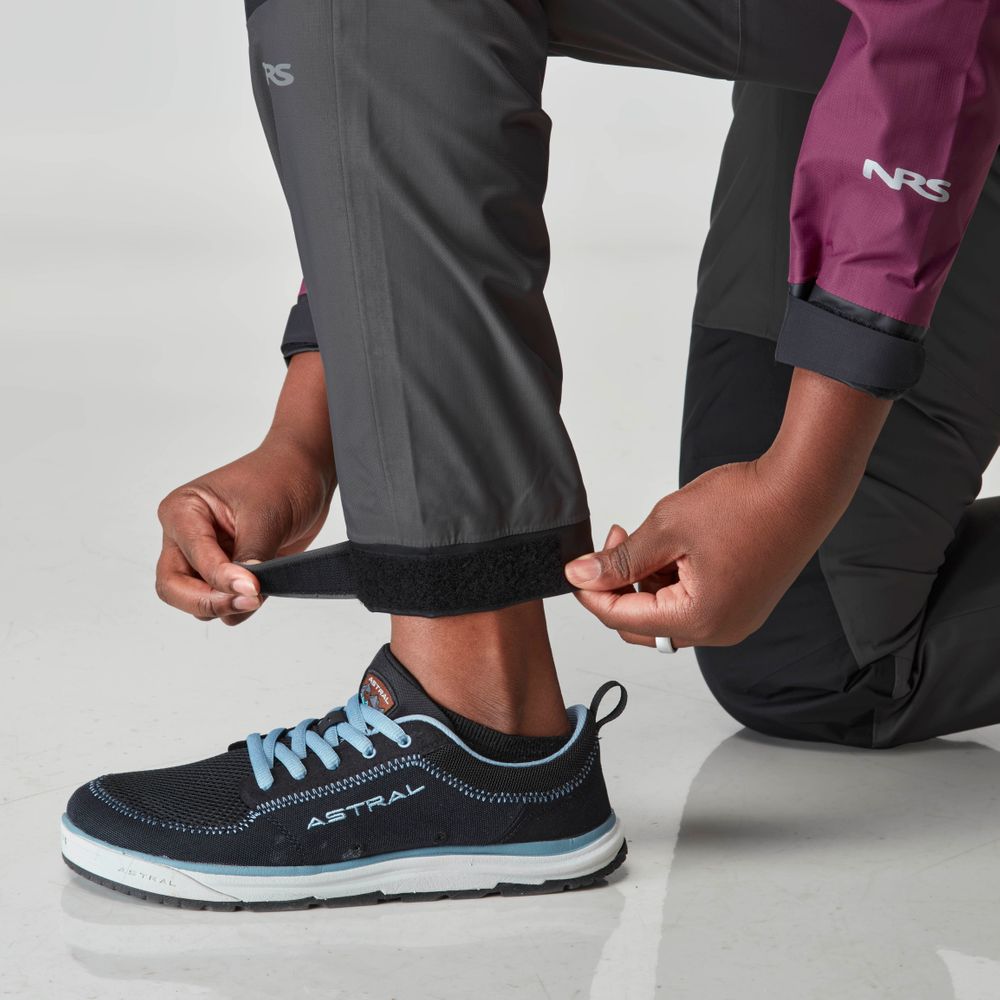 Adjusting the Velcro strap on NRS's Endurance Splash Pant in dark gray HyproTex, these women's waterproof pants pair perfectly with a maroon jacket and black sneakers with blue accents, all against a light-colored floor.