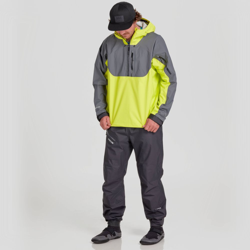 A man stands against a plain background in the NRS High Tide Jacket—a bright yellow and gray waterproof splash jacket with an adjustable hood—paired with matching gray pants and black shoes.