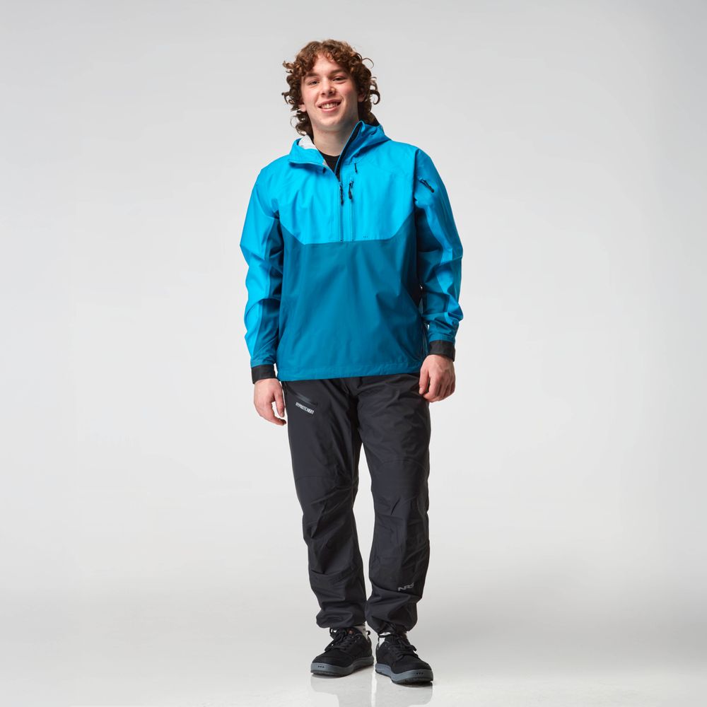 A man wearing the NRS High Tide Jacket - Men's in blue and teal, featuring a waterproof design with an adjustable hood, smiles at the camera, paired with black pants.