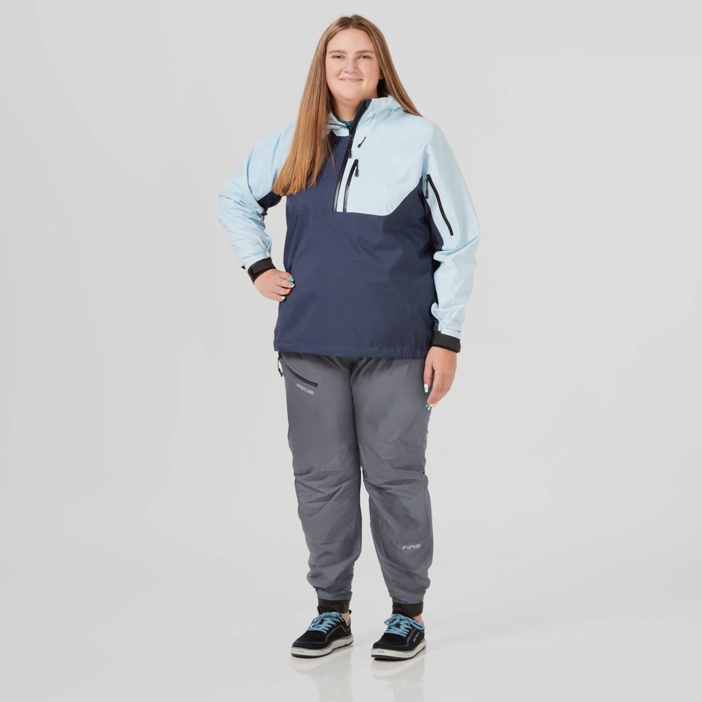 A person stands confidently against a plain background, wearing the NRS High Tide Jacket - Women's in blue and gray. They smile with one hand on their hip, highlighting the paddler's cut for ultimate adventure comfort.