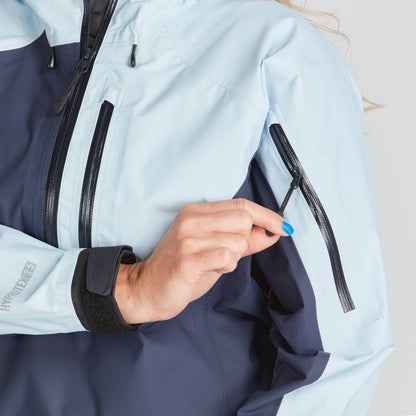 A person zips a pocket on the sleeve of an NRS High Tide Jacket - Women's in light blue and dark blue, wearing a smartwatch.