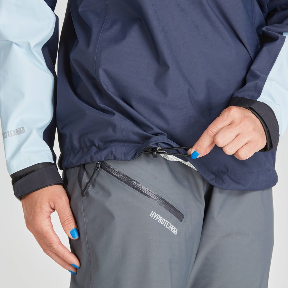 A person adjusts the drawstring on the NRS High Tide Jacket - Women's in dark blue, paired with gray waterproof pants featuring a visible zip pocket.