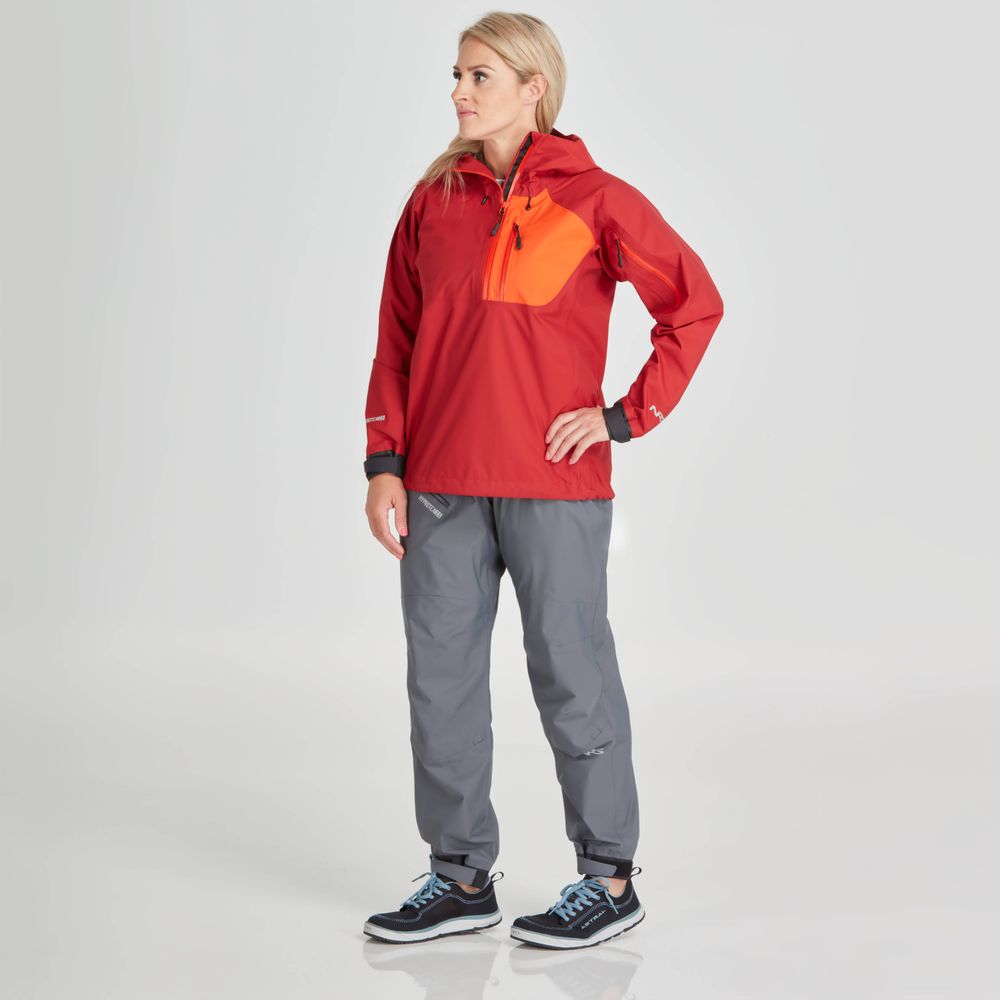 A woman confidently poses in an NRS High Tide Jacket in red and orange, paired with gray pants and black sneakers, against a plain background.