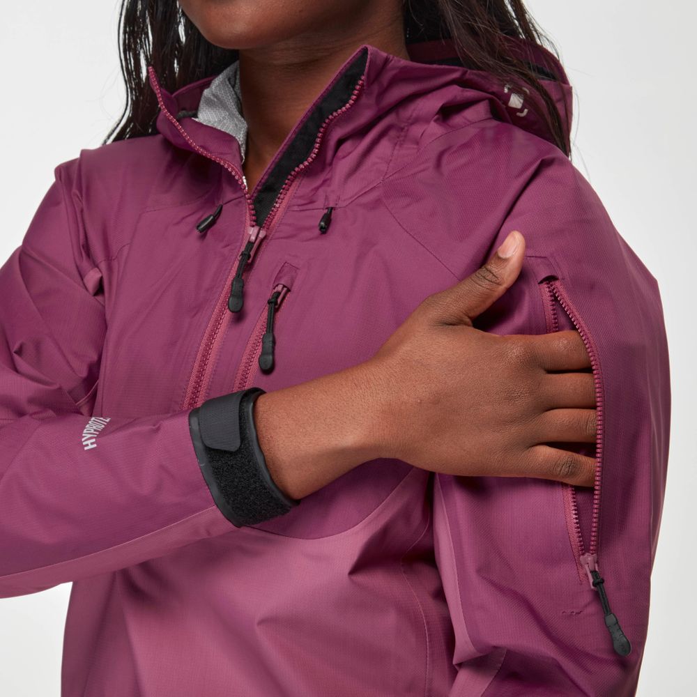 A person wearing the maroon High Tide Jacket - Women's by NRS adjusts the sleeve pocket zipper.