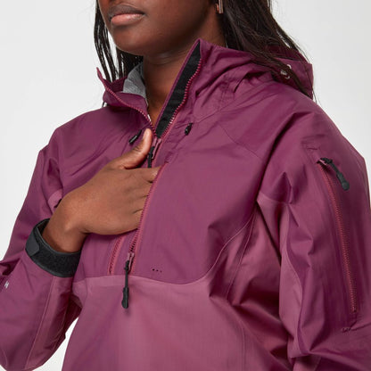 A person wearing the NRS High Tide Jacket - Women's in magenta, featuring a hood and zippered pockets, holds the front zipper with one hand.
