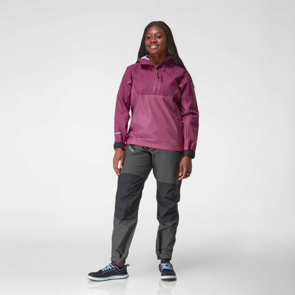 A person smiles against a plain background, modeling the NRS High Tide Jacket for women—a chic purple splash jacket featuring an adjustable hood—perfectly matched with gray pants.