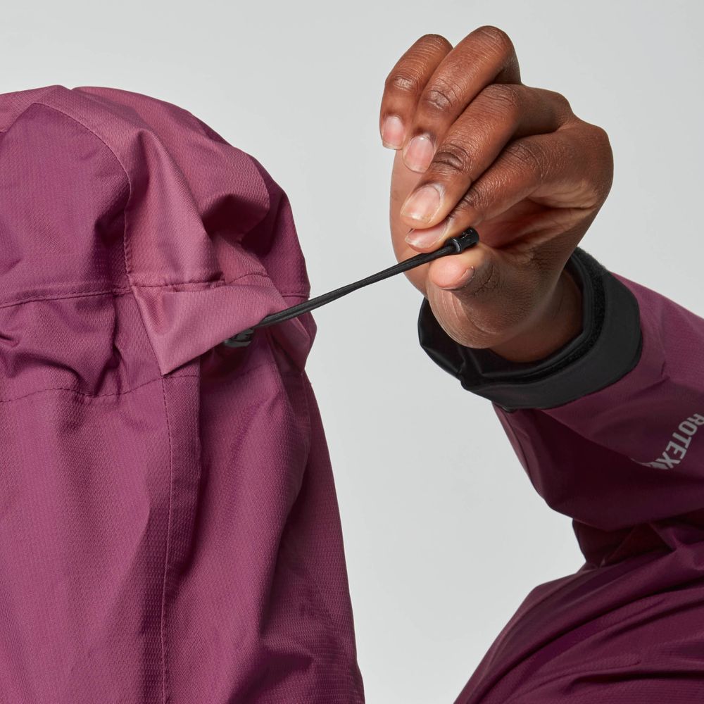A woman tightens the hood drawcord of the NRS High Tide Jacket, ensuring a secure fit before stepping out by the water.
