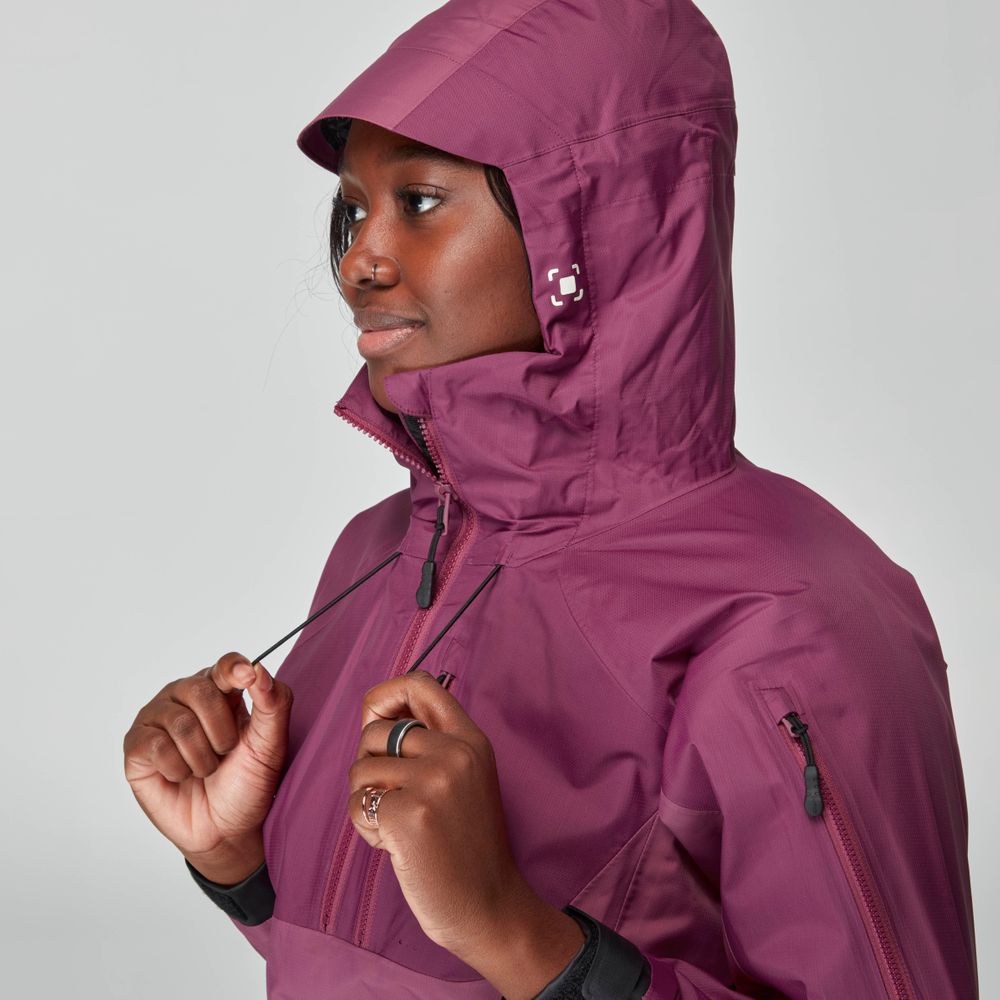A person dressed in the purple NRS High Tide Jacket for women, with a hood and waterproof design, adjusts the drawstrings while facing left against a gray background.