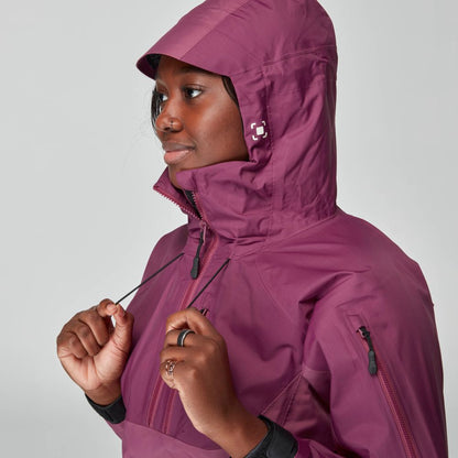 A person dressed in the purple NRS High Tide Jacket for women, with a hood and waterproof design, adjusts the drawstrings while facing left against a gray background.