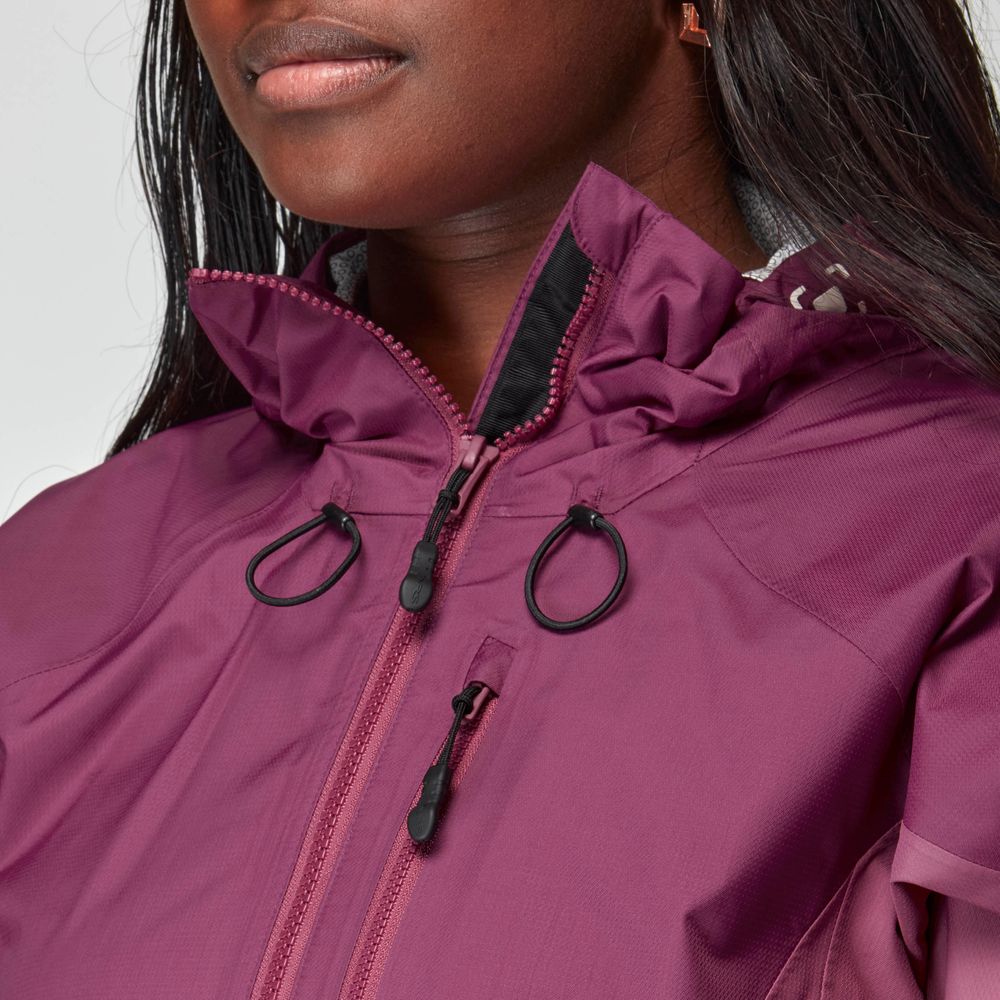 A woman wears the NRS High Tide Jacket—a purple waterproof splash jacket with a high collar, black zipper pulls, and drawstring cords—shown from the shoulders up.