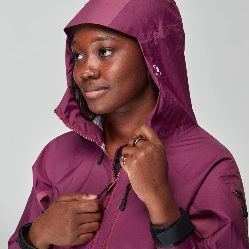 A woman wearing the NRS High Tide Jacket - Women's in magenta adjusts the zipper while looking to the side, set against a plain light background.