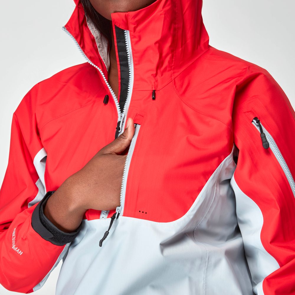 She's wearing the NRS High Tide Jacket in red and gray, confidently gripping the zipper of its waterproof design with a high collar.