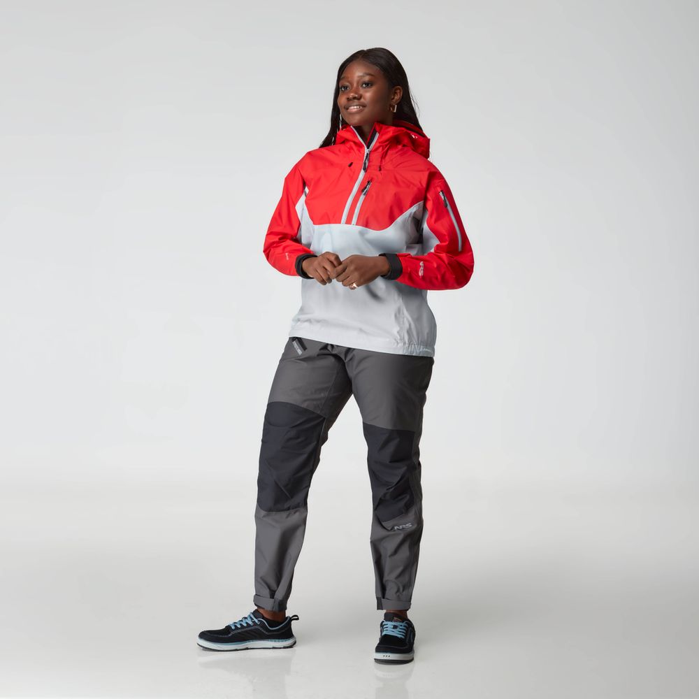 In a studio, a woman models the NRS High Tide Jacket - Women's in red and gray, paired with gray pants and black sneakers featuring blue soles. The jacket includes an adjustable hood.