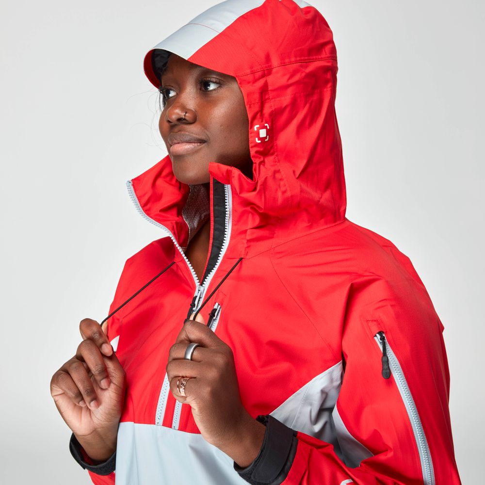 A woman adjusts the hood's drawstrings on a red and gray waterproof High Tide Jacket by NRS, ensuring breathable protection against a plain background.